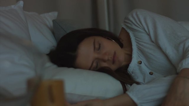 Young woman going to bed indoors interior close up 4k handheld camera. Attractive girl sleeping lying white linen fabric natural light evening routine. Insomnia healthy lifestyle pills drugs problems
