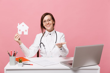 Female doctor sit at desk work on computer with medical document hold house in hospital isolated on pastel pink wall background. Woman in medical gown glasses stethoscope. Healthcare medicine concept.