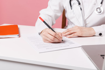 Cropped female doctor siting at desk working filling out medical documents in hospital isolated on pastel pink wall background indoors. Woman in medical gown stethoscope. Healthcare medicine concept.