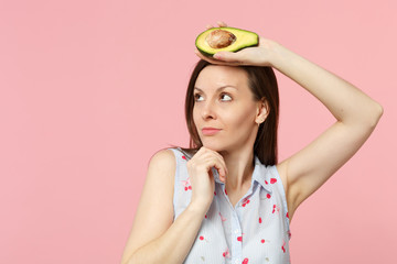 Pensive young woman looking aside put hand prop up on chin holding fresh ripe green avocado fruit isolated on pink pastel background. People vivid lifestyle relax vacation concept. Mock up copy space.