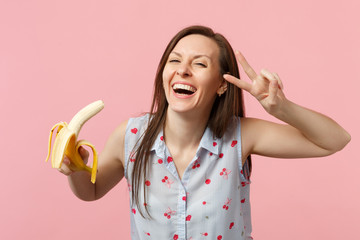 Laughing young woman in summer clothes showing victory sign holding fresh ripe banana fruit isolated on pink pastel wall background. People vivid lifestyle, relax vacation concept. Mock up copy space.