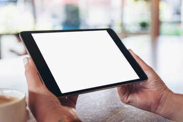 Mockup image of hands holding black tablet pc with blank white screen horizontally with coffee cup on wooden table
