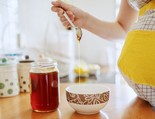 Close up of pregnant Caucasian woman in apron putting honey from jar in bowl while standing in...