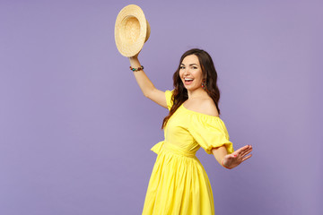 Side view of cheerful young woman in yellow dress holding summer hat spreading hands isolated on pastel violet wall background in studio. People sincere emotions lifestyle concept. Mock up copy space.