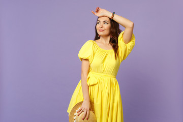 Smiling young woman in yellow dress holding summer hat, putting hand on head, looking aside isolated on pastel violet wall background. People sincere emotions, lifestyle concept. Mock up copy space.