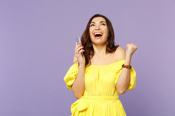 Happy young woman in summer dress using mobile phone, typing sms message, doing winner gesture, looking up isolated on violet background. People sincere emotions lifestyle concept. Mock up copy space.