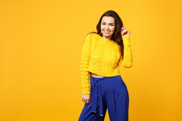 Portrait of smiling young woman in sweater, blue trousers looking camera rising hand isolated on yellow orange wall background in studio. People sincere emotions lifestyle concept. Mock up copy space.