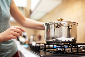 Beautiful young woman housewife prepairing dinner, hold in hands big steel saucepan, standing it on gas-stove.