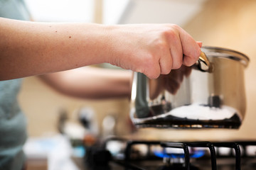 Beautiful young woman housewife prepairing dinner, hold in hands big steel saucepan, standing it on gas-stove.