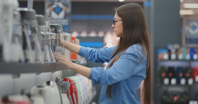 Young beautiful brunette woman chooses an electric kettle to buy. Holds a device in his hands, studies the price tags and the characteristics and design of the model