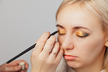 Fototapeta premium Visagiste with a brush in one hand putting makeup on the eyes of the model, the other hand pulls the skin of the eyelid with gold and green shadows in east style.