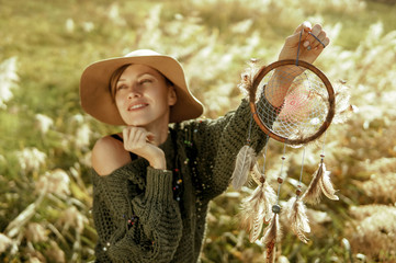 Beautiful woman wearing hat and boho outfit holding a dream catcher in a sunny morning. Live in a...