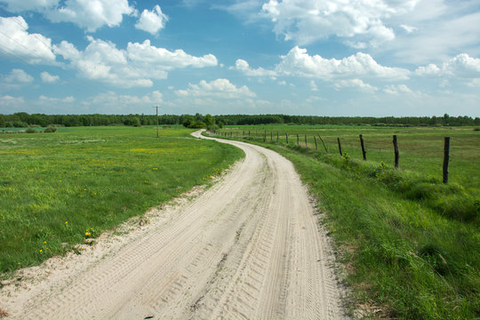Sandy winding road through green pastures, wooden posts in the fence