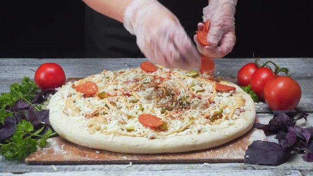 Chef's hands in silicone gloves adding the pieces of saveloy on a pizza lying near tomatoes, parsley and basil. Frame. Delicious pizza preparation