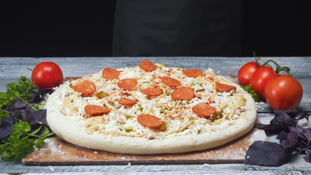 Close-up of pizza covered with pieces of saveloy and cheese lying on a wooden board near tomatoes, parsley and basil. Frame. Delicious pizza preparation