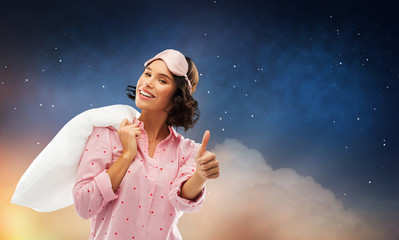 people and bedtime concept - happy young woman in pajama and eye sleeping mask with pillow over shoulder showing thumbs up over starry night sky background