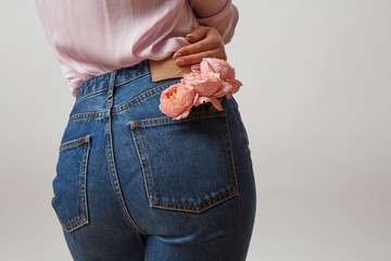Attractive woman's butt in a blue jeans and fresh roses living coral color in a back pocket on a light gray background, place for text. Congratulation card.
