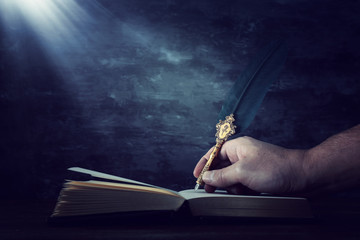 Old feather quill ink pen with inkwell and old books over wooden desk in front of black wall...