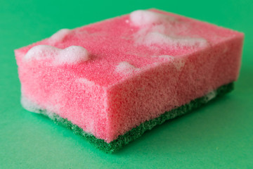 Red kitchen sponge in the foam from dishwashing detergent. The item is on a green background. Minimalistic image is suitable as a background.