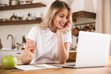 Image of modern blond woman studying on laptop and writing down notes while sitting at wooden table...