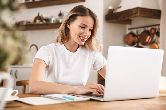 Image of european blond woman 20s wearing white t-shirt studying on laptop and writing down notes in apartment