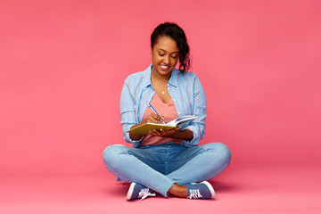 education, high school and people concept - happy african american young student woman writing to diary or notebook sitting on floor over pink background