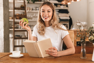 Portrait of beautiful blond woman reading book and eating green apple while sitting in cozy cafe...