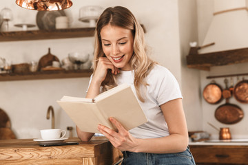 Portrait of young blond woman reading book while standing in stylish wooden kitchen at home