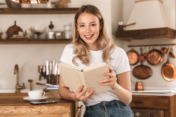 Portrait of relaxed blond woman reading book while standing in stylish wooden kitchen at home