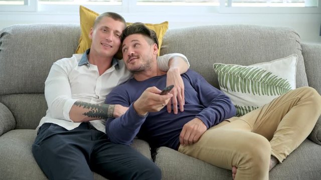 Young gay male couple relaxing on couch in living room watching tv. Caucasian, european men sitting on sofa. Switch channel and talking. Happy gay friendship, relationship, lifestyle concept. 