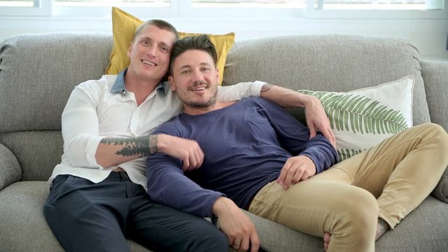 Young gay male couple relaxing on couch in living room watching tv. Caucasian, european men sitting on sofa. Laughing together. Happy gay friendship, relationship, lifestyle concept. 