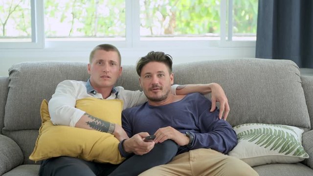 Young gay male couple relaxing on couch in living room watching tv. Caucasian, european men sitting on sofa. Scare mood. Happy gay friendship, relationship, lifestyle concept. 
