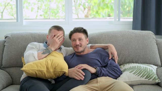 Young gay male couple relaxing on couch in living room watching tv. Caucasian, european men sitting on sofa. Jump scare. Happy gay friendship, relationship, lifestyle concept. 