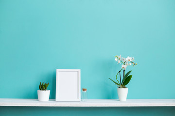 Modern room decoration with Picture frame mockup. White shelf against pastel turquoise wall with potted orchid and succulent plant.