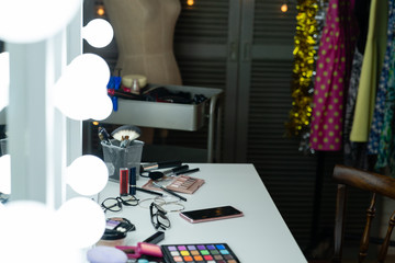 Stylish clothes and make up products in dressing room in program company backstage. cosmetics and cellphone on table by mirror with lights in empty room interior nobody. dress hang on clothing rack