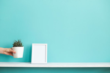 Modern room decoration with Picture frame mockup. White shelf against pastel turquoise wall with hand putting down potted succulent plant.