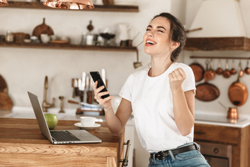 Beautiful young student girl sitting indoors using laptop computer chatting by mobile phone.