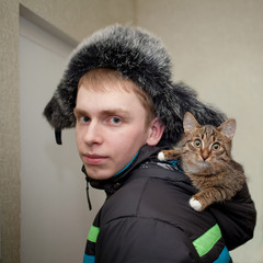 young man with red hair wearing a hat with earflaps with fur with a tabby color kitten in the hood...