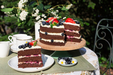 Slice of layered chocolate cake decorated with flowers and berries on the garden table.