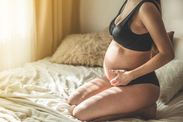 Attractive pregnant woman is sitting in bed and holding her belly. Last months of pregnancy