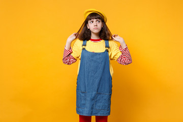 Portrait of pensive girl teenager in french beret, denim sundress holding hair, looking up isolated on yellow wall background in studio. People sincere emotions, lifestyle concept. Mock up copy space.