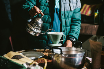 Man wearing coat and making pour-over coffee with alternative method called Dripping. Coffee grinder, coffee stand and pour-over on a wooden table.