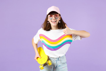 Smiling teen girl in vivid clothes, heart eyeglasses hold yellow skateboard, showing thumb up isolated on violet pastel wall background. People sincere emotions, lifestyle concept. Mock up copy space.