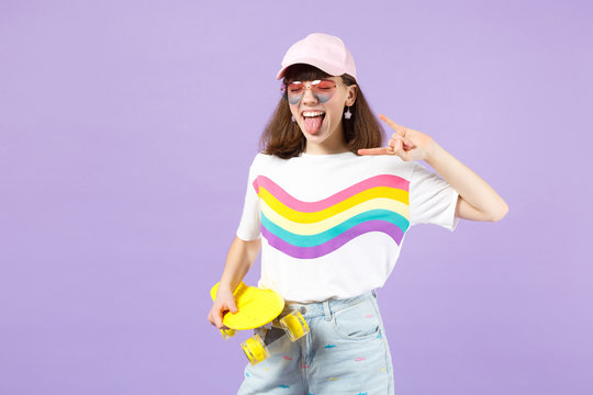 Crazy teen girl with skateboard depicting heavy metal rock sign, showing tongue keeping eyes closed isolated on violet pastel background. People sincere emotions lifestyle concept. Mock up copy space.