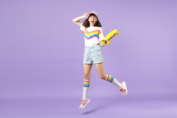 Portrait of laughing teen girl in vivid clothes holding yellow skateboard, having fun jumping isolated on violet pastel wall background. People sincere emotions, lifestyle concept. Mock up copy space.