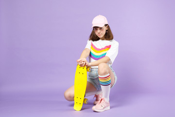 Charming teen girl in vivid clothes sitting hold yellow skateboard, looking camera isolated on violet pastel wall background in studio. People sincere emotions, lifestyle concept. Mock up copy space.
