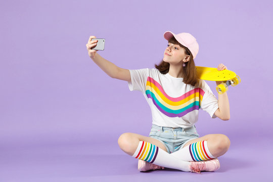 Beautiful teen girl in vivid clothes holding yellow skateboard, doing selfie shot on mobile phone isolated on violet pastel background. People sincere emotions, lifestyle concept. Mock up copy space.