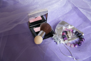 Blush and mirror, brush, ring and bracelet on a purple fabric background. Women's cosmetics and accessories.