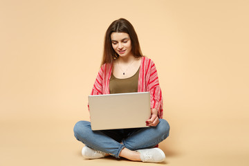 Naklejka premium Portrait of beautiful young woman in casual clothes sitting, using laptop pc computer isolated on pastel beige wall background in studio. People sincere emotions lifestyle concept. Mock up copy space.
