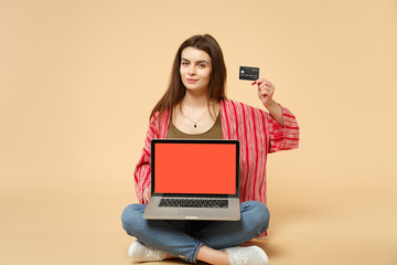 Beautiful young woman sitting, holding credit bank card, laptop pc computer with blank empty screen isolated on pastel beige background. People sincere emotions, lifestyle concept. Mock up copy space.